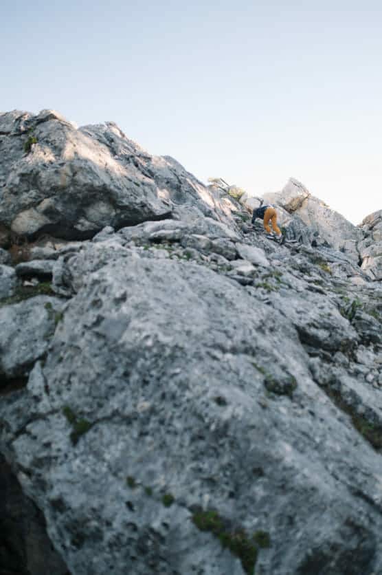 A person wearing an orange jacket climbs over large, rugged gray rocks on a mountainside under a clear blue sky. by Justin Salem Meyer