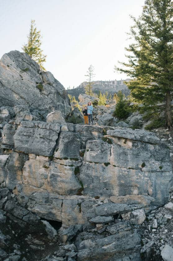 Two people stand on a rocky outcrop surrounded by large gray boulders and tall pine trees, with mountains visible in the background under a clear sky. by Justin Salem Meyer