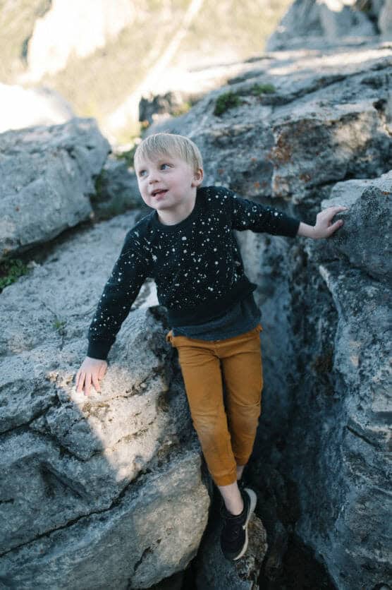 A young child with blonde hair, wearing a black speckled sweater and mustard yellow pants, climbs between large gray rocks outdoors on a sunny day. by Justin Salem Meyer