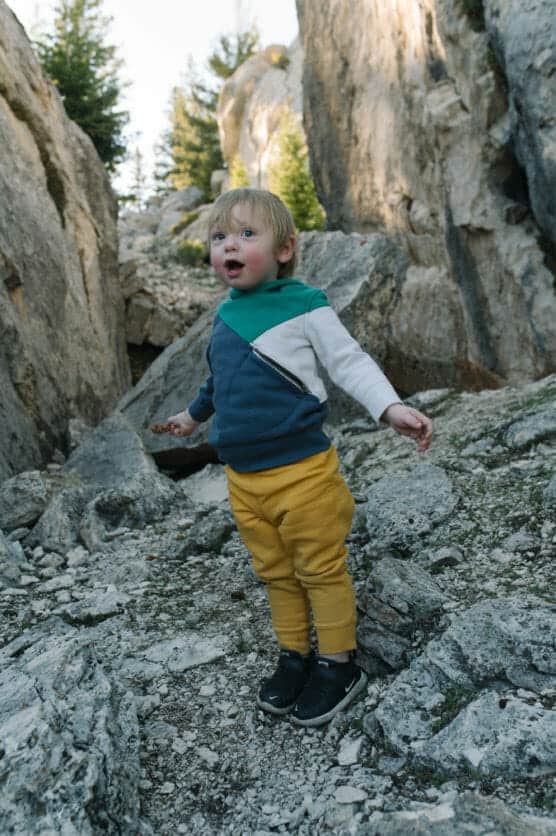 A young child with blond hair stands on rocky ground in a mountainous area, wearing a blue, green, and white jacket with yellow pants and black shoes, with trees and large rocks in the background. by Justin Salem Meyer