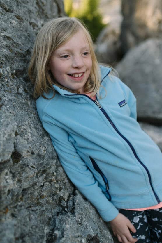 A smiling child with blonde hair and a light blue jacket leans against a large rock outdoors, with sunlight highlighting their face. by Justin Salem Meyer