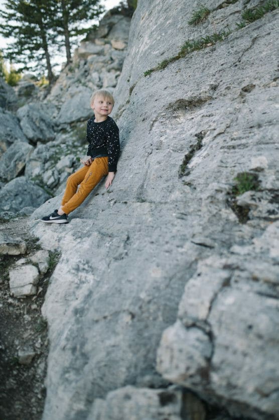 A young child with blonde hair, wearing a black long-sleeve shirt with white specks and mustard-colored pants, sits and smiles on a large sloped gray rock outdoors, surrounded by rocky terrain and trees. by Justin Salem Meyer