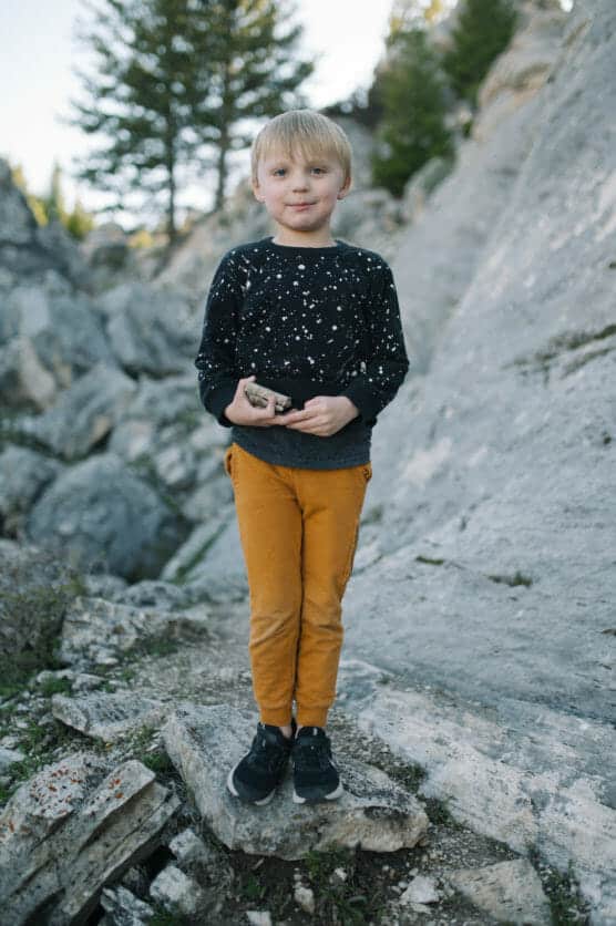 A young boy with blonde hair stands on rocky terrain, wearing a black and white speckled sweater, mustard yellow pants, and black shoes. Pine trees and large rocks are visible in the background. by Justin Salem Meyer