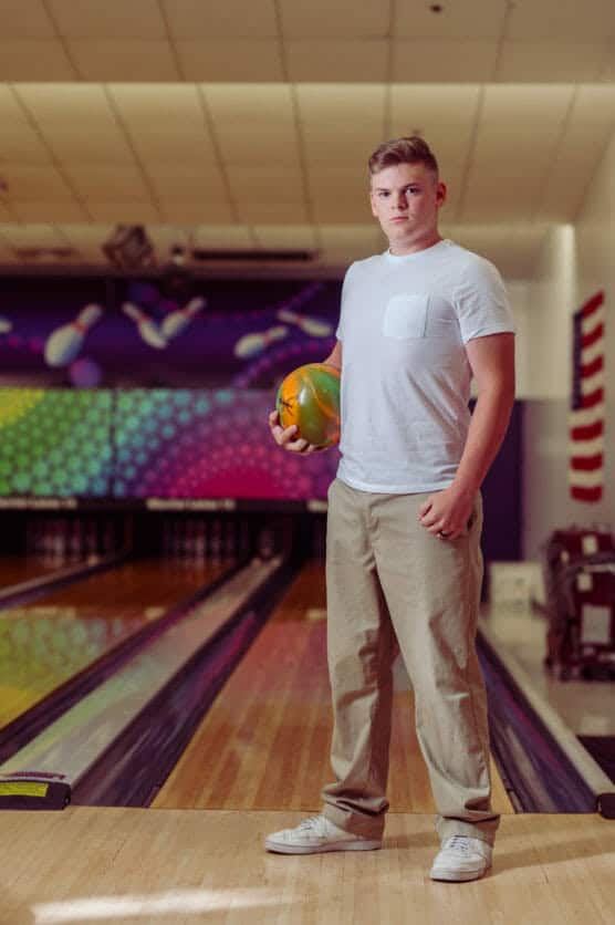 A young man in a white t-shirt and beige pants stands at the start of a bowling lane, holding a bowling ball. The bowling alley features colorful wall art and an American flag in the background. by Justin Salem Meyer