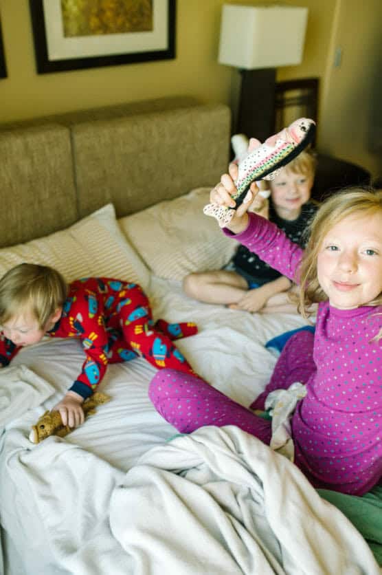 Three young children in pajamas play on a bed. One girl in purple polka-dot pajamas smiles and holds up a toy fish, while the other two children play with stuffed animals next to her. by Justin Salem Meyer