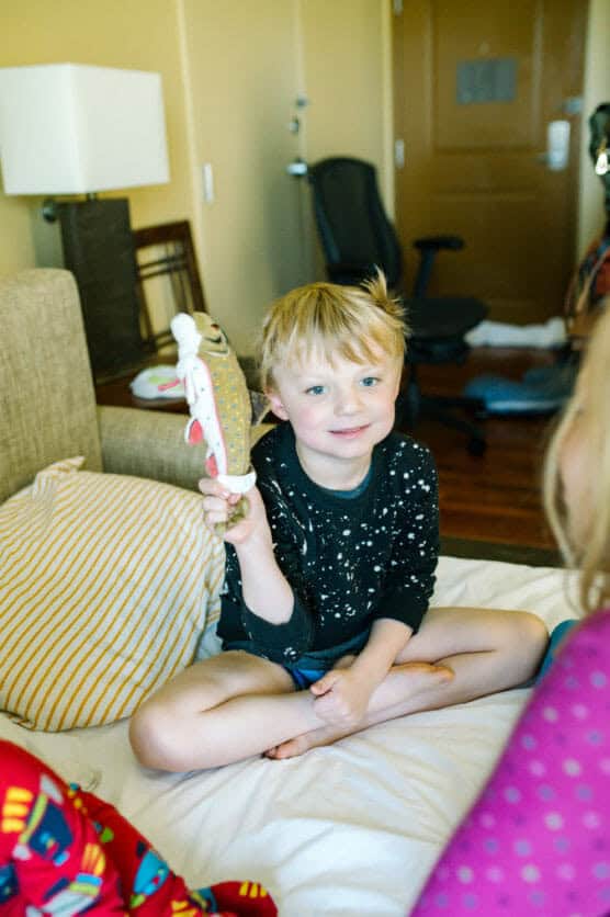 A young boy with blonde hair sits cross-legged on a bed, smiling and holding up a plush toy. He is wearing a black shirt with white speckles. Another child in colorful pajamas is partially visible in the foreground. by Justin Salem Meyer