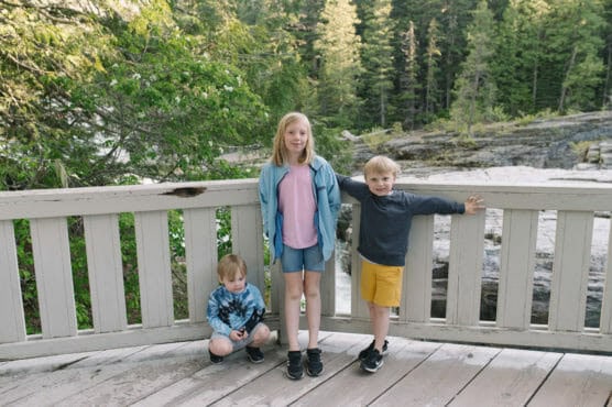 Three young children stand and pose on a wooden deck with a white railing, surrounded by trees and a rocky river in the background. Two boys stand by the rail, and a girl stands in the middle, all dressed in casual summer clothes. by Justin Salem Meyer