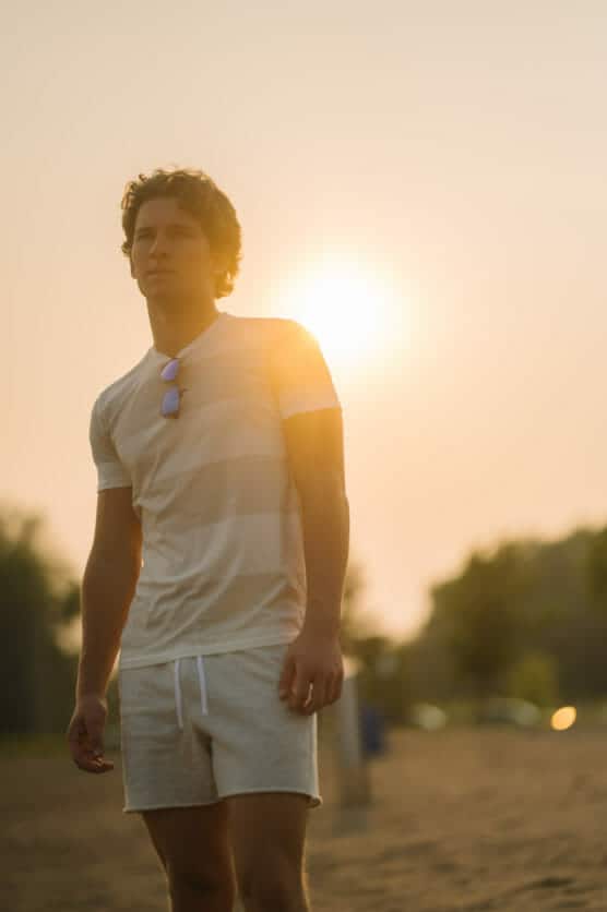 A young man wearing a striped t-shirt and light shorts stands on a sandy beach at sunset, with the sun glowing behind him and sunglasses hanging from his shirt collar. by Justin Salem Meyer