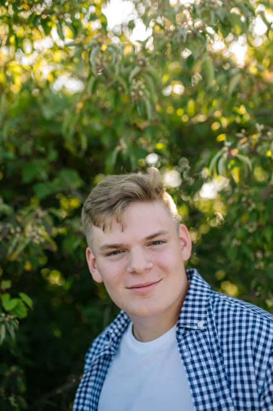A young man with short blond hair, wearing a white T-shirt and a blue checkered shirt, stands outdoors in front of leafy green bushes with sunlight filtering through the branches. by Justin Salem Meyer