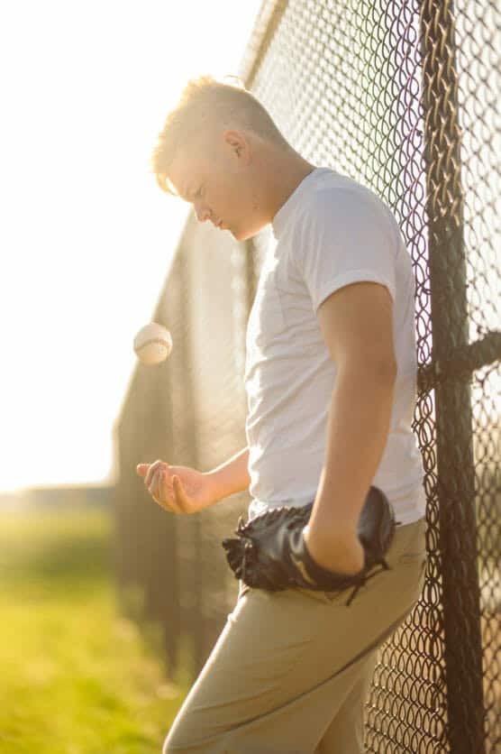 A young person in a white shirt and khaki pants leans against a chain-link fence, tossing a baseball in the air with one hand and holding a glove in the other, bathed in warm sunlight. by Justin Salem Meyer