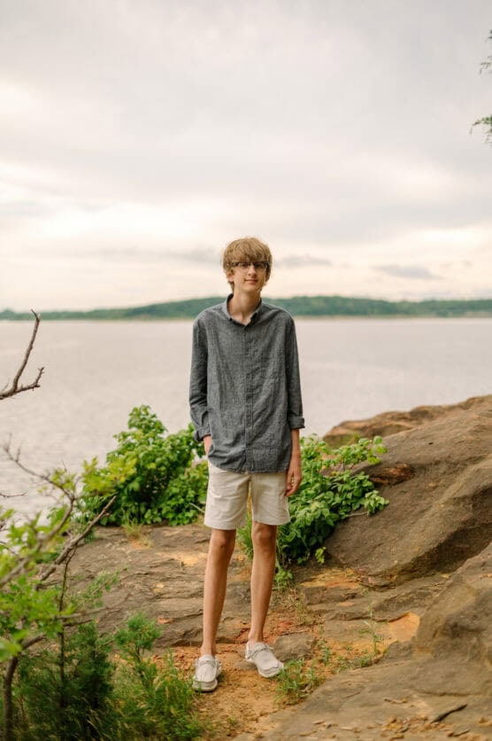 A teenage boy with light brown hair, glasses, a gray button-up shirt, and beige shorts stands on a rocky outcrop near a body of water, with greenery and a distant shoreline visible in the background. by Justin Salem Meyer