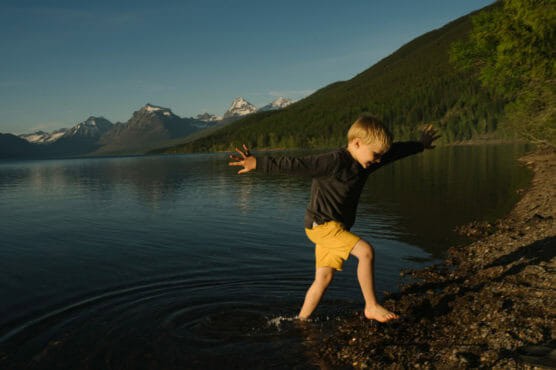 A young boy in a dark shirt and yellow shorts splashes barefoot at the edge of a calm lake, surrounded by mountains and forest under a clear sky. by Justin Salem Meyer
