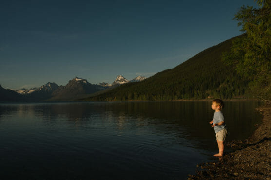 A small child stands at the edge of a calm lake surrounded by pine-covered mountains and snow-capped peaks under a clear sky, bathed in warm sunlight. by Justin Salem Meyer