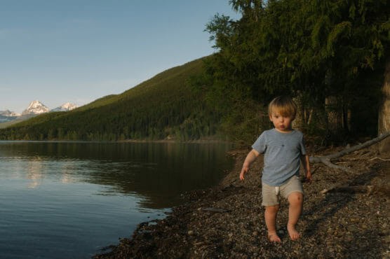 A young child walks barefoot on a rocky lakeshore, with calm water, forested hills, and snow-capped mountains in the background under a clear sky. by Justin Salem Meyer