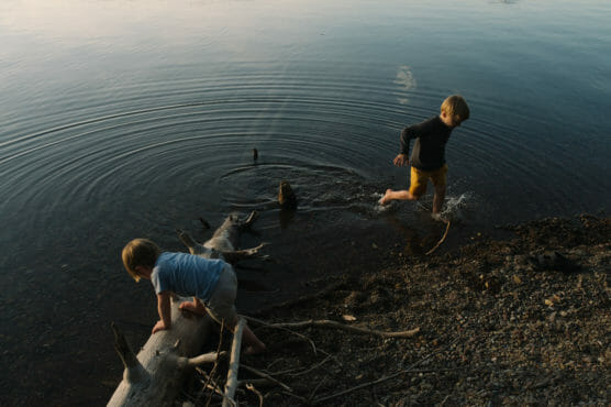 Two young children play at the edge of a calm lake. One walks on a fallen log in the water while the other splashes nearby, creating gentle ripples. The shoreline is rocky and scattered with driftwood. by Justin Salem Meyer