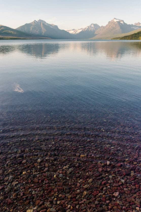 Clear, calm lake with smooth, multicolored pebbles visible through the water in the foreground. Snow-capped mountains and pine forests surround the lake under a bright sky in the background. by Justin Salem Meyer