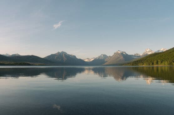 Calm lake reflecting snow-capped mountains and a clear blue sky, with a forested shoreline on the right side. by Justin Salem Meyer