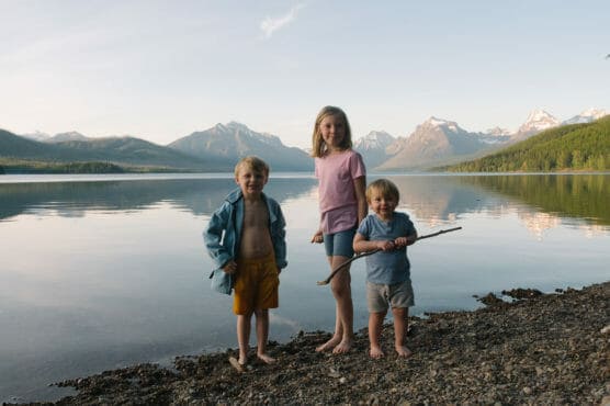 Three young children stand barefoot on a rocky lakeshore with mountains and trees reflected in the calm water behind them. One child holds a stick. The setting appears peaceful and natural. by Justin Salem Meyer