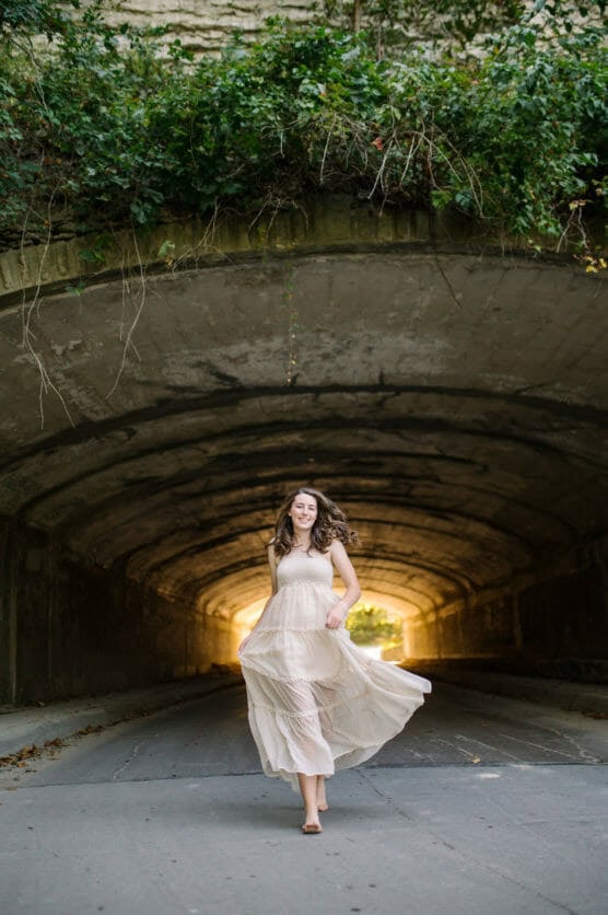 A woman in a flowing, light-colored dress walks barefoot under a concrete tunnel with greenery growing above, sunlight glowing at the tunnel’s far end. by Justin Salem Meyer