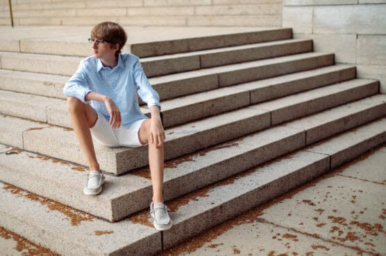 A young person with short hair, glasses, a blue shirt, and white shorts sits on wide stone steps, looking to the side. Brown leaves are scattered on the steps around them. by Justin Salem Meyer