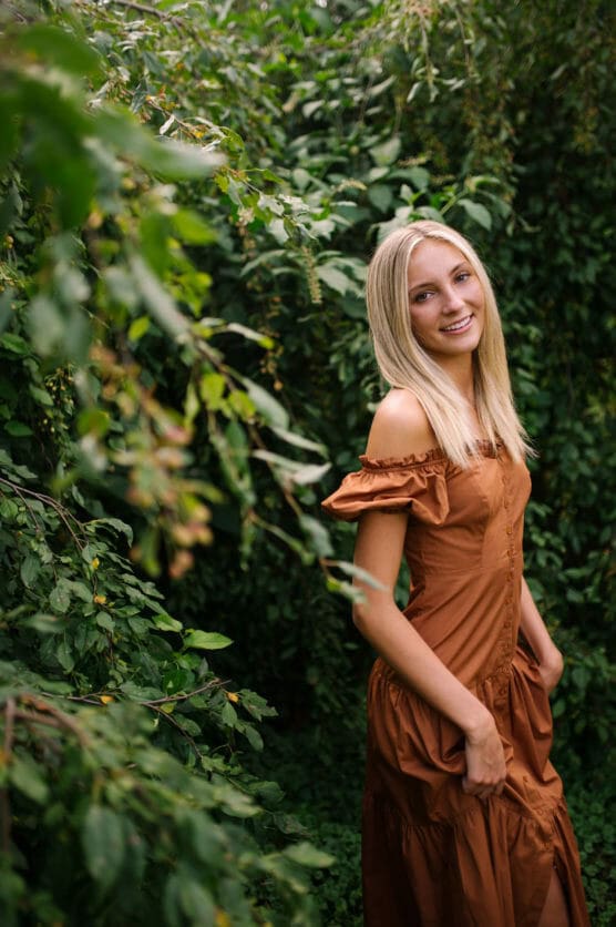 A young woman with straight blonde hair, wearing an off-the-shoulder brown dress, stands smiling among lush green foliage in an outdoor setting. by Justin Salem Meyer
