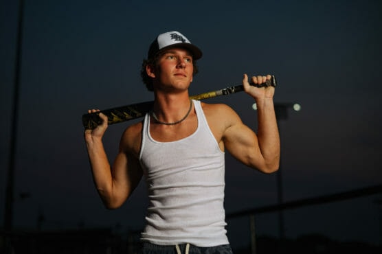 A young man wearing a white tank top, necklace, and baseball cap rests a baseball bat across his shoulders while standing outdoors at dusk. by Justin Salem Meyer