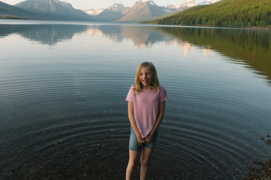 A young girl in a pink shirt and denim shorts stands in shallow water at the edge of a calm lake, with forested hills and snow-capped mountains reflected in the water behind her. by Justin Salem Meyer