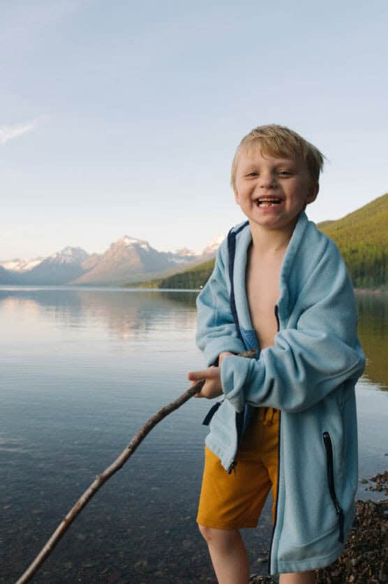A smiling young boy in a blue jacket and yellow shorts stands by a calm lake, holding a stick, with snow-capped mountains and green trees in the background. by Justin Salem Meyer