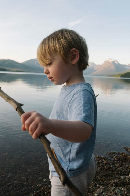 A young boy with blond hair, wearing a light blue shirt, stands by a calm lake holding a stick, with mountains and a clear sky in the background. by Justin Salem Meyer