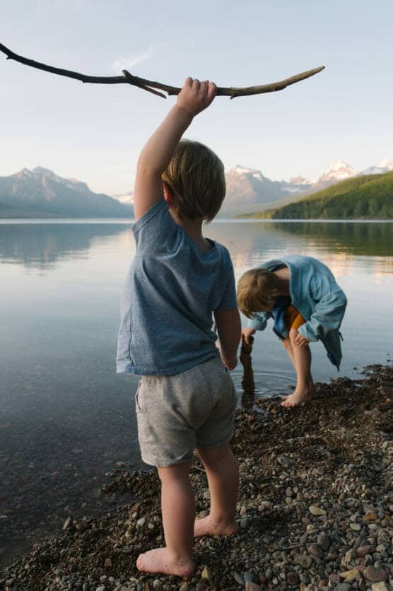 Two young children play barefoot on a rocky lakeshore; one holds up a stick while the other squats near the water. Snow-capped mountains and trees are visible in the background. by Justin Salem Meyer