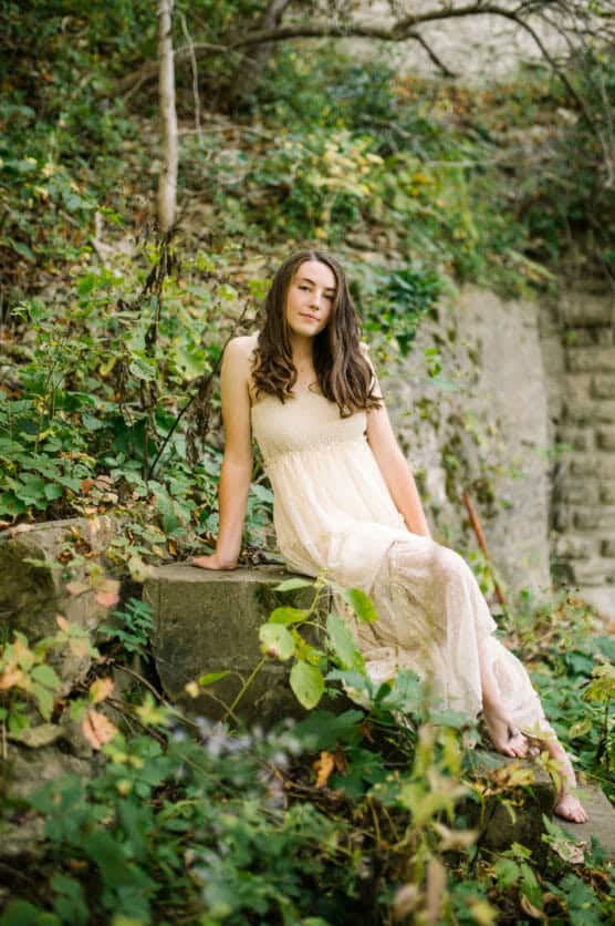 A young woman in a light, sleeveless dress sits on a stone ledge surrounded by lush greenery and foliage, with a stone wall in the background. She looks relaxed and serene in the natural outdoor setting. by Justin Salem Meyer