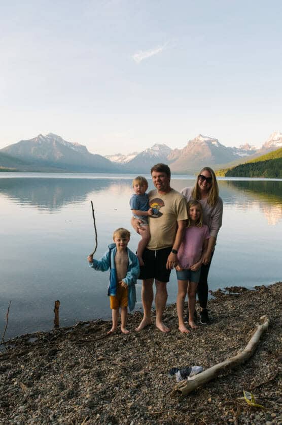 A family of five stands together on a pebbled lakeshore with mountains and trees in the background. The father holds a toddler, while the other children and the mother stand nearby, smiling on a calm, sunny day. by Justin Salem Meyer