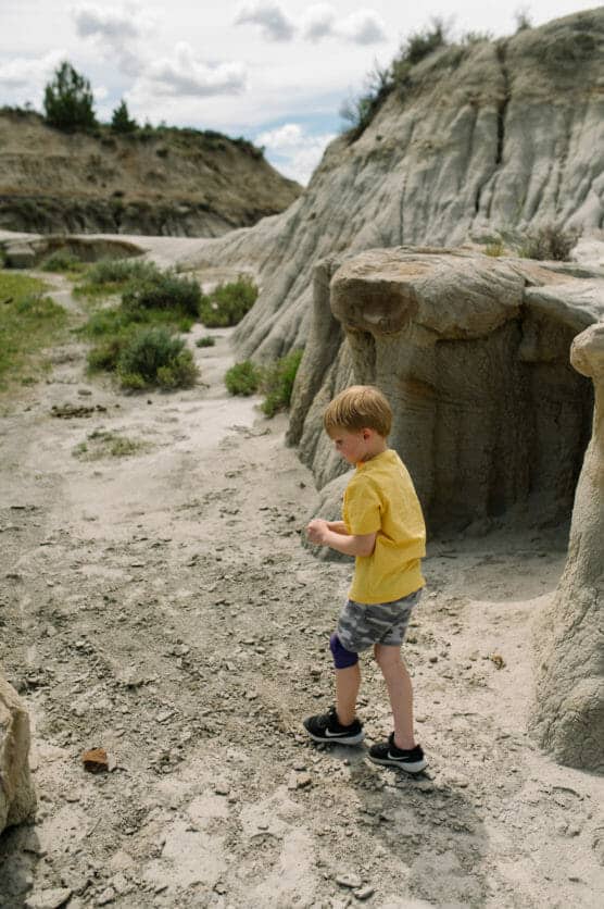 A young boy wearing a yellow shirt, shorts, and sneakers walks through a rocky, sandy landscape with eroded hills and sparse vegetation under a cloudy sky. by Justin Salem Meyer