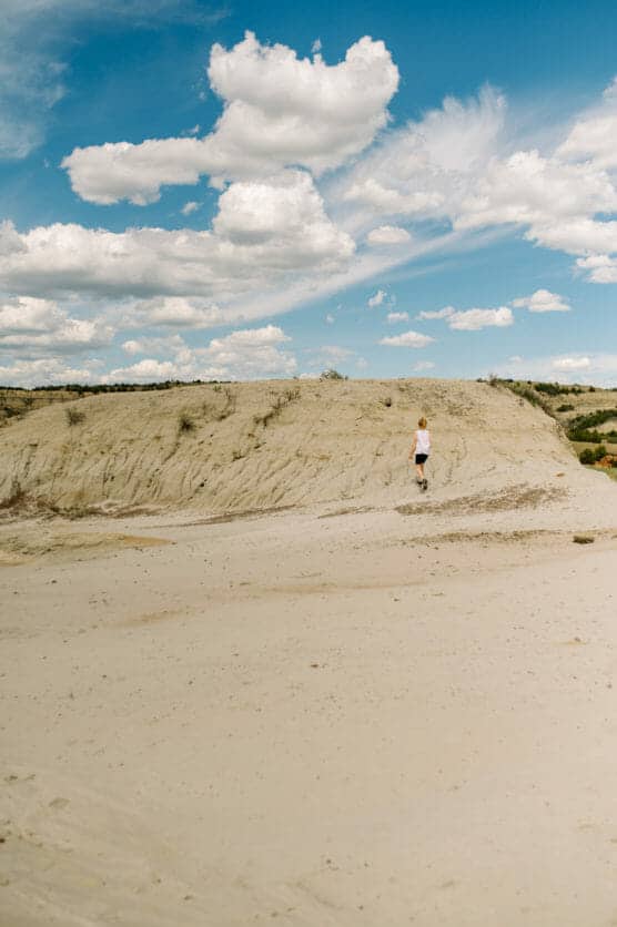 A person wearing shorts and a white shirt walks alone across a sandy, barren landscape under a blue sky with scattered, fluffy white clouds. by Justin Salem Meyer