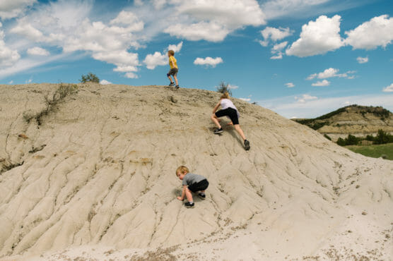 Three children climb up a sandy, rocky hill under a blue sky with scattered clouds. One child is near the top, while the other two are midway up the slope. The landscape is open with distant hills and greenery. by Justin Salem Meyer