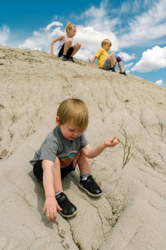 Three young children sit and play on a sandy, rocky hill under a bright blue sky with clouds. The closest child holds a small stick, while the other two watch from higher up the hill. by Justin Salem Meyer