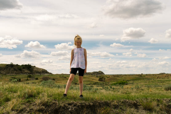 A young girl stands on grassy ground in front of rolling hills under a partly cloudy sky, looking down with her hands at her sides, wearing a sleeveless top, black shorts, and black shoes. by Justin Salem Meyer