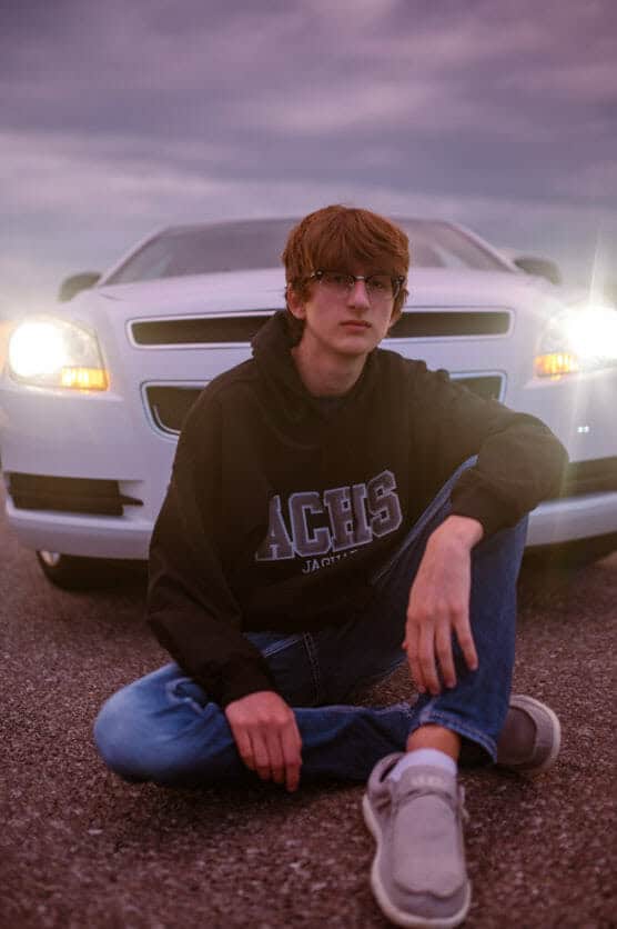 A young man with brown hair and glasses sits cross-legged on the pavement in front of a white car with its headlights on, wearing a black hoodie, jeans, and gray shoes, under a cloudy sky. by Justin Salem Meyer
