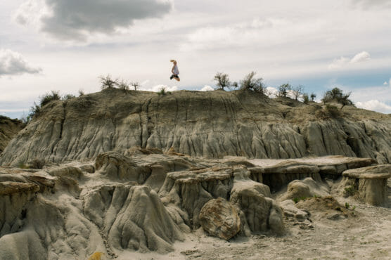 A person jumps in midair above rocky, eroded badlands with sparse vegetation under a cloudy sky. The landscape features layered rock formations and a dramatic, rugged terrain. by Justin Salem Meyer