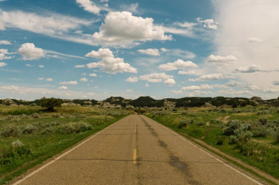 A straight, empty road stretches into the distance through grassy fields and low hills under a blue sky filled with scattered white clouds. by Justin Salem Meyer