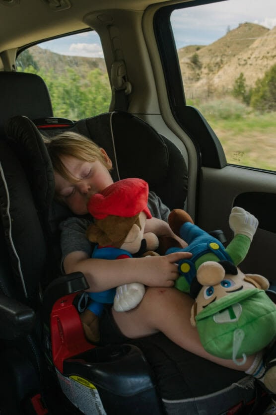 A young child sleeps in a car seat, holding plush toys of Mario and Luigi, while sunlight streams through the car window and a scenic landscape is visible outside. by Justin Salem Meyer