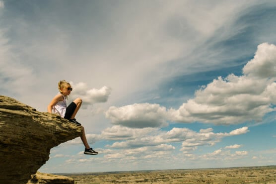 A person sits on the edge of a rocky cliff under a partly cloudy sky, overlooking a vast landscape. They wear sunglasses, a white tank top, and black shorts and sneakers. by Justin Salem Meyer
