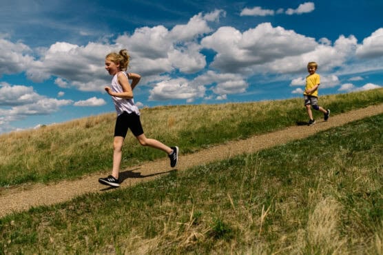Two children run along a dirt path through a grassy field under a bright blue sky with scattered clouds. The girl in front wears a white shirt and black shorts, while the boy behind her wears a yellow shirt and gray shorts. by Justin Salem Meyer