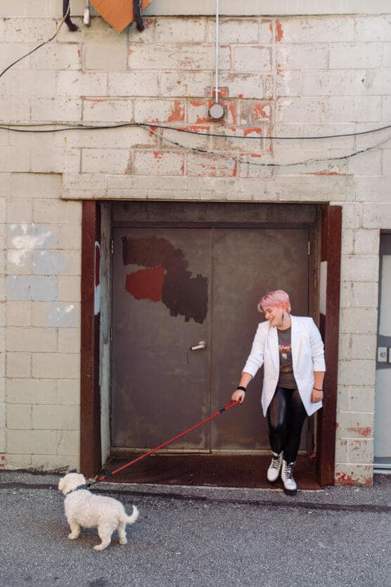 A person with pink hair, dressed in a white coat and black pants, stands in a doorway holding a red leash attached to a small white dog. The background is a weathered brick wall with peeling paint. by Justin Salem Meyer
