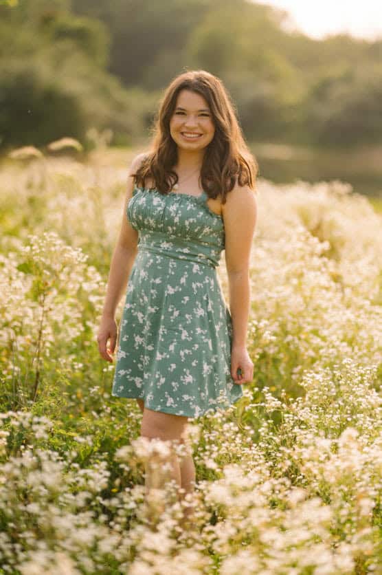 A young woman with long brown hair, wearing a green floral dress, stands smiling in a sunlit field of white wildflowers, with blurred greenery in the background. by Justin Salem Meyer