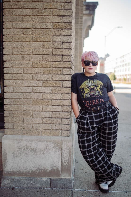Person with short pink hair and sunglasses leans against a brick wall, wearing a black Queen band t-shirt, black and white plaid wide-leg pants, and white shoes on a city sidewalk. by Justin Salem Meyer