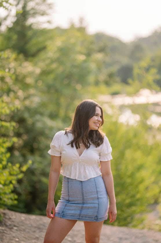 A woman with long brown hair, wearing a white blouse and a light blue denim skirt, stands outdoors in a sunlit, green natural setting, smiling and looking to the side. by Justin Salem Meyer