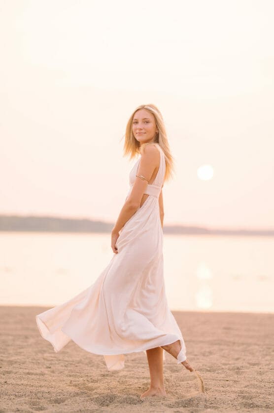 A young woman in a flowing white dress stands barefoot on a sandy beach at sunset, looking at the camera with a gentle smile. The water and sun are visible in the background, creating a warm, serene atmosphere. by Justin Salem Meyer