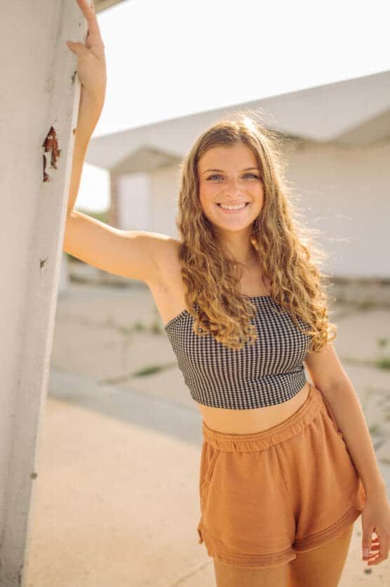 A young woman with long curly hair smiles at the camera while leaning against a white wall outside. She is wearing a checkered crop top and orange shorts on a sunny day. by Justin Salem Meyer