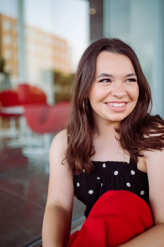 A young woman with long brown hair, wearing a black polka dot top, sits smiling near a glass wall with red chairs and buildings blurred in the background. by Justin Salem Meyer